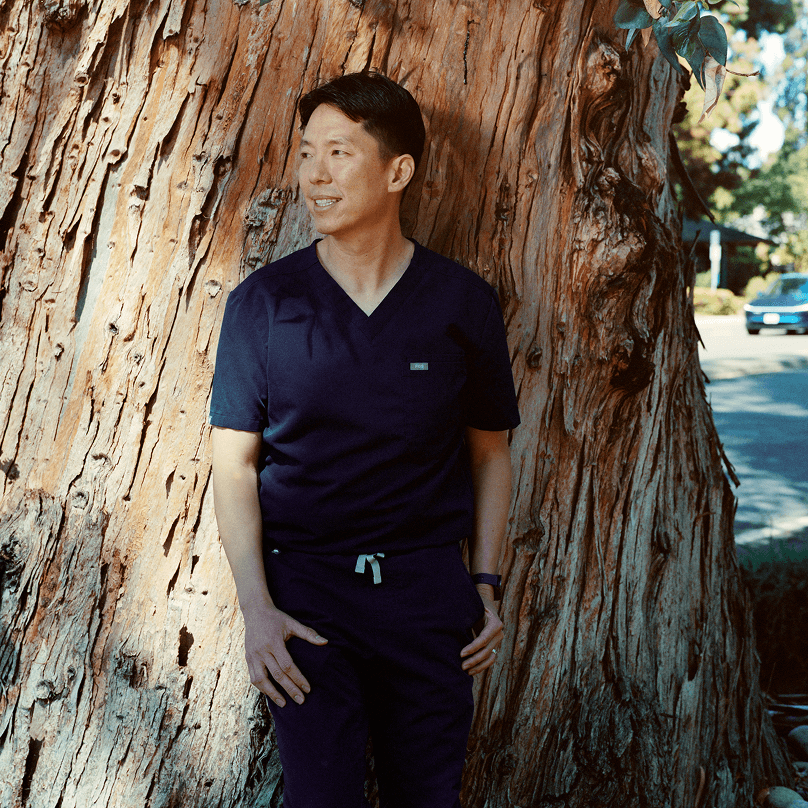 A man wearing dark blue scrubs stands outdoors, leaning gently against the rough, deeply textured bark of a large tree trunk. He is looking thoughtfully off-camera to the side, illuminated by warm sunlight.